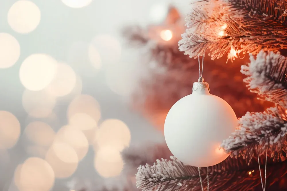 Weiße Christbaumkugel hängt an schneebedecktem Zweig eines beleuchteten Weihnachtsbaums mit warmem Bokeh-Hintergrund